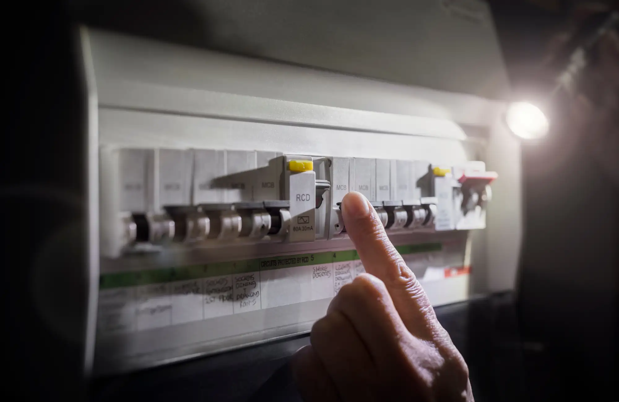 An electrician Brooklyn is about to press the yellow test button on a circuit breaker switch inside an NY electrical panel, with labels and switches clearly visible under bright light.