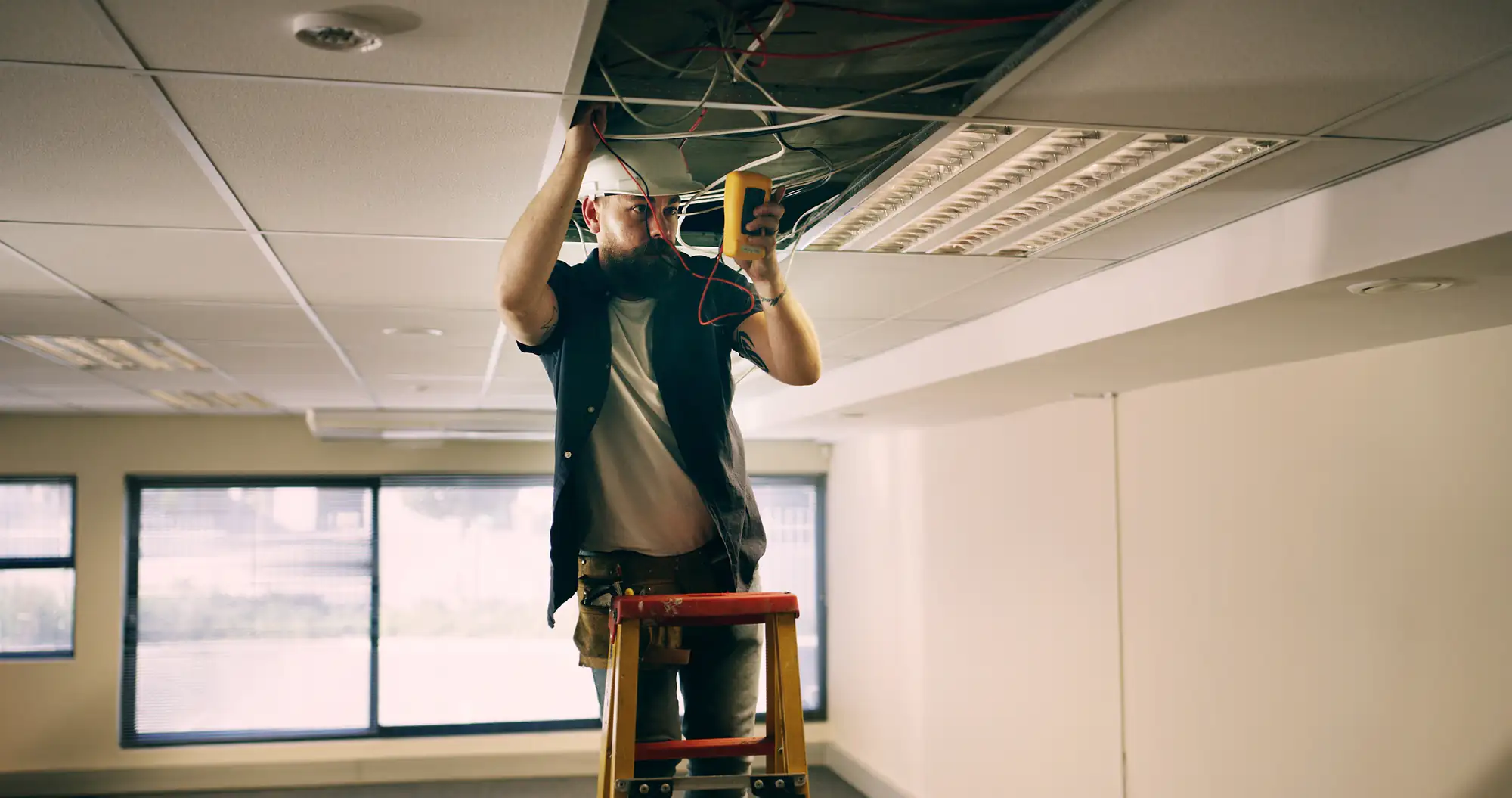An electrician in Brooklyn, NY, wearing a hard hat stands on a ladder and uses electrical testing equipment to inspect wiring inside an open ceiling panel in an office setting.