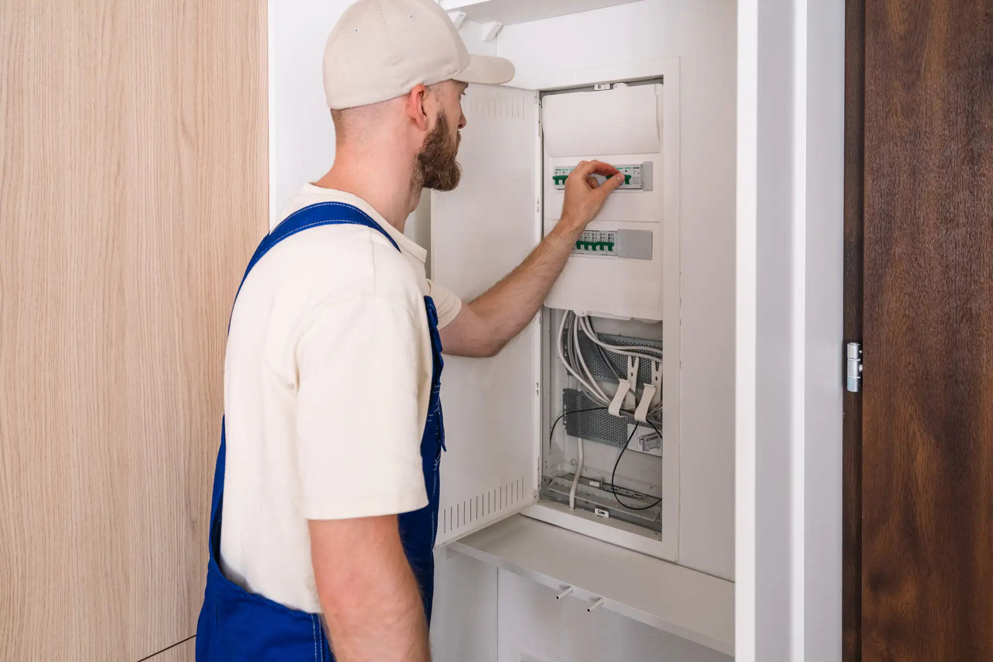 An electrician Brooklyn in NY, wearing a cap and blue overalls, is carefully adjusting switches inside a wall-mounted electrical panel with exposed wiring. The open panel door reveals his focused work on the wiring.