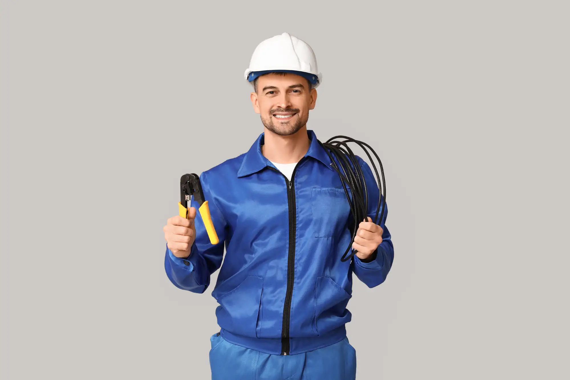 A smiling electrician Brooklyn, NY, wearing a blue uniform and white hard hat holds wire strippers and a bundle of black cables, standing against a plain gray background.