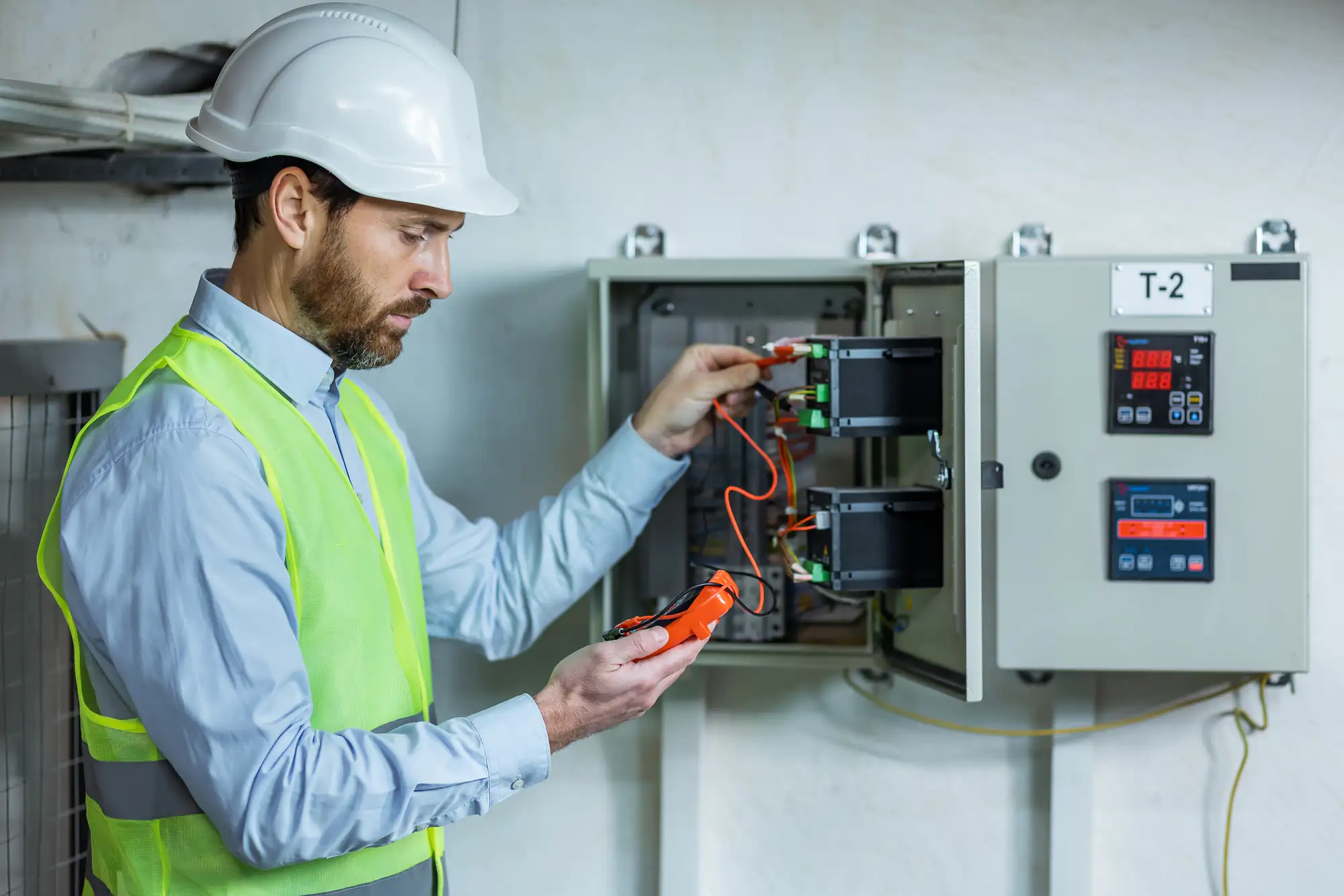 A Brooklyn electrician wearing a white hard hat and yellow safety vest uses a multimeter to check electrical connections inside an open control panel mounted on a wall in NY.