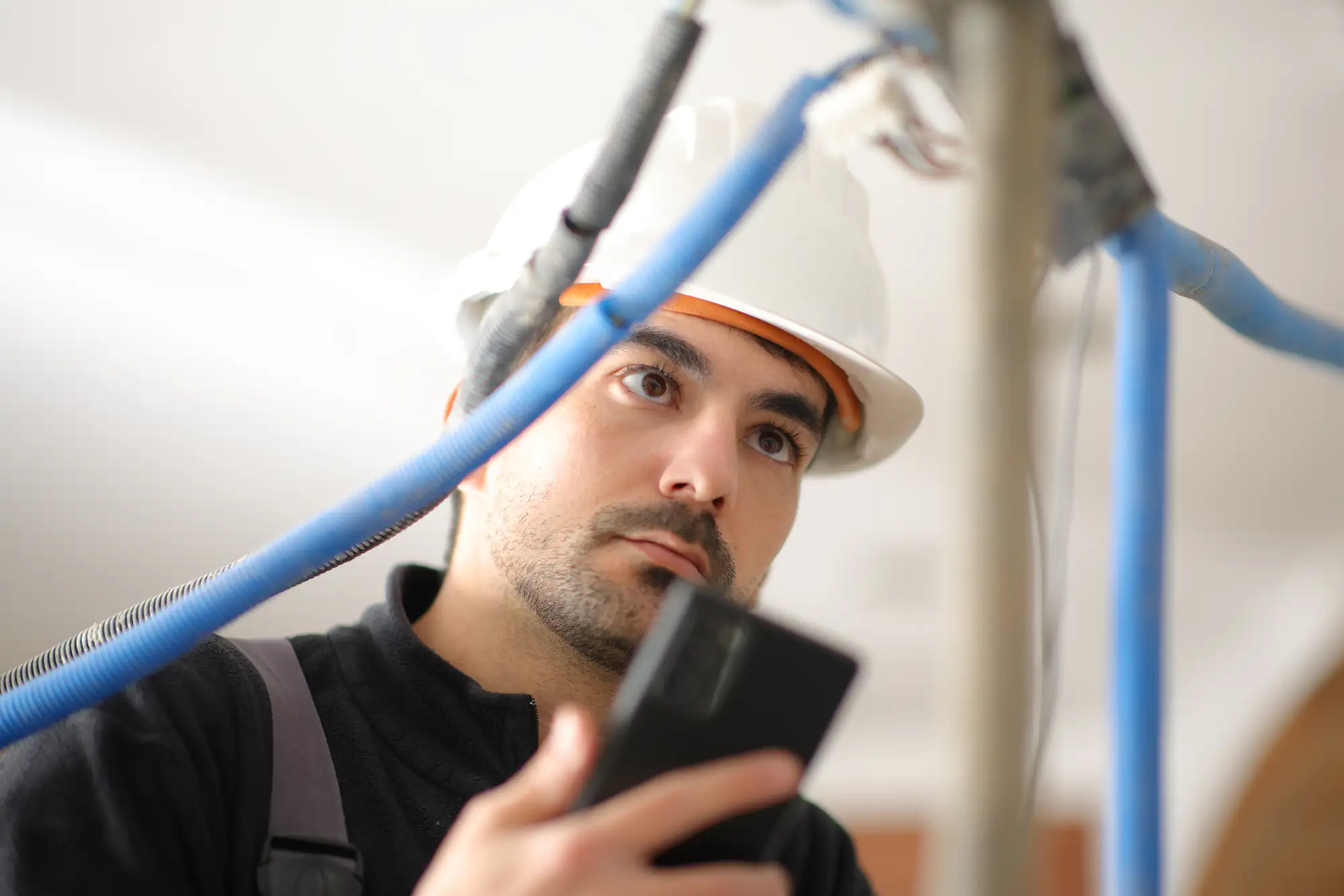 A construction worker, possibly an electrician Brooklyn, wearing a white hard hat examines blue cables while holding a smartphone or device in NY, appearing focused on the inspection.