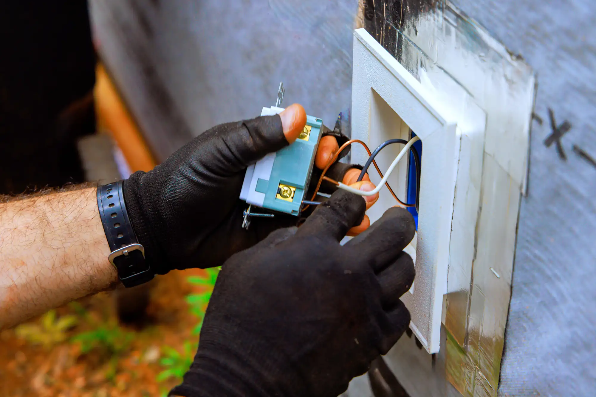 An electrician Brooklyn in NY, wearing black gloves, installs or repairs a wall electrical outlet, connecting wires to the terminals on the back of the socket, with tools and materials visible around the work area.