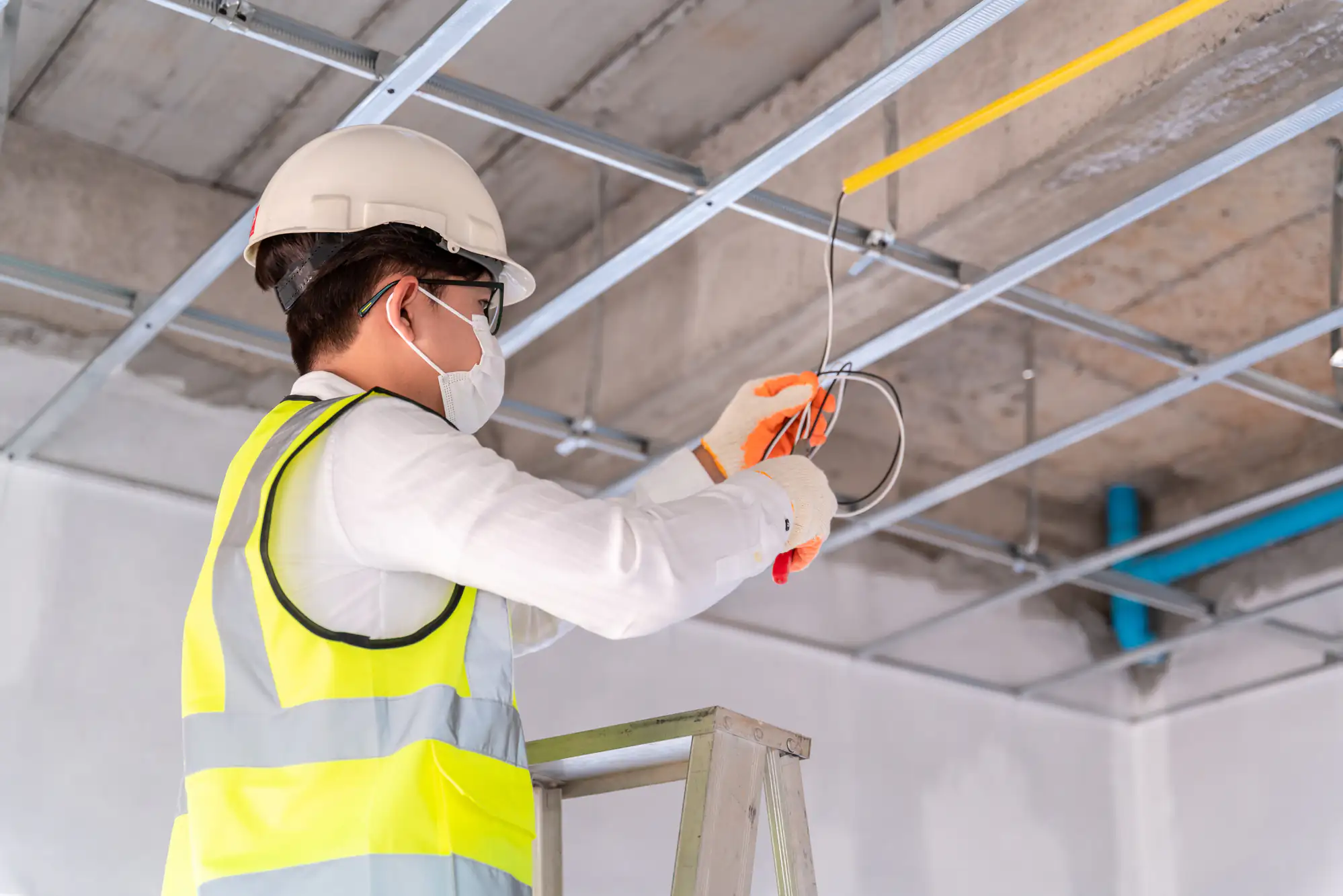 A construction worker, possibly an electrician Brooklyn, wearing a hard hat, safety vest, gloves, and face mask stands on a ladder while installing electrical wiring on a NY building&rsquo;s ceiling framework.