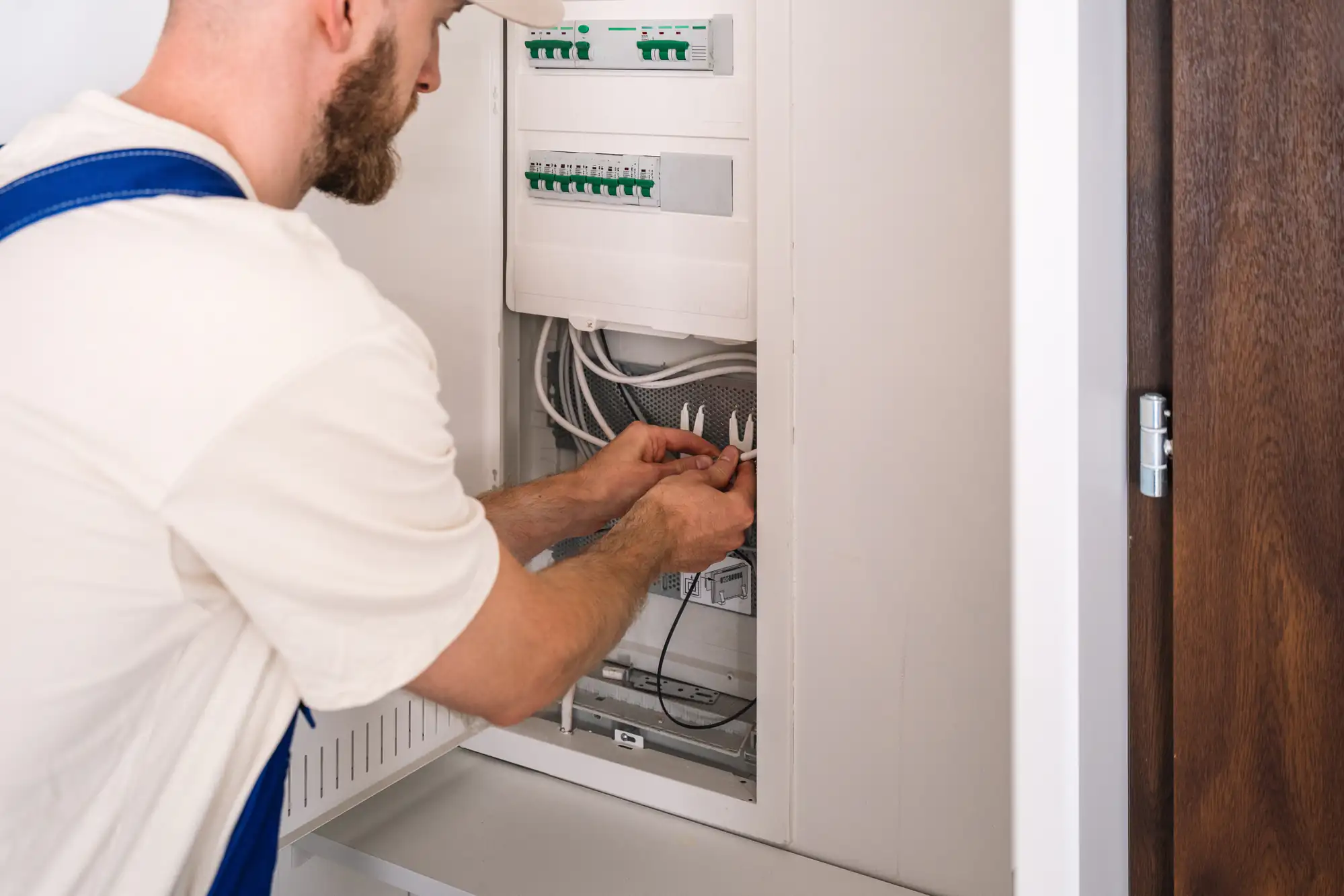 An electrician in Brooklyn, NY, wearing a white shirt and blue overalls, works on an open electrical panel, connecting or inspecting wires inside a utility cabinet next to a brown wooden door.