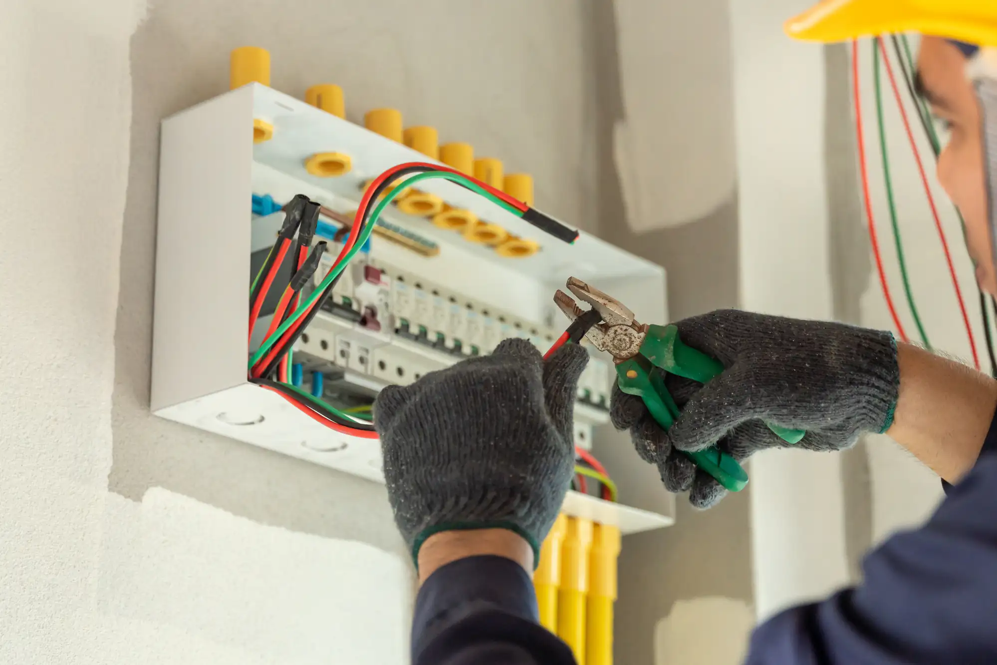 An electrician Brooklyn, NY, wearing gloves uses pliers to work on electrical wires inside an open circuit breaker panel mounted on a wall. Yellow conduit pipes are visible below the panel.
