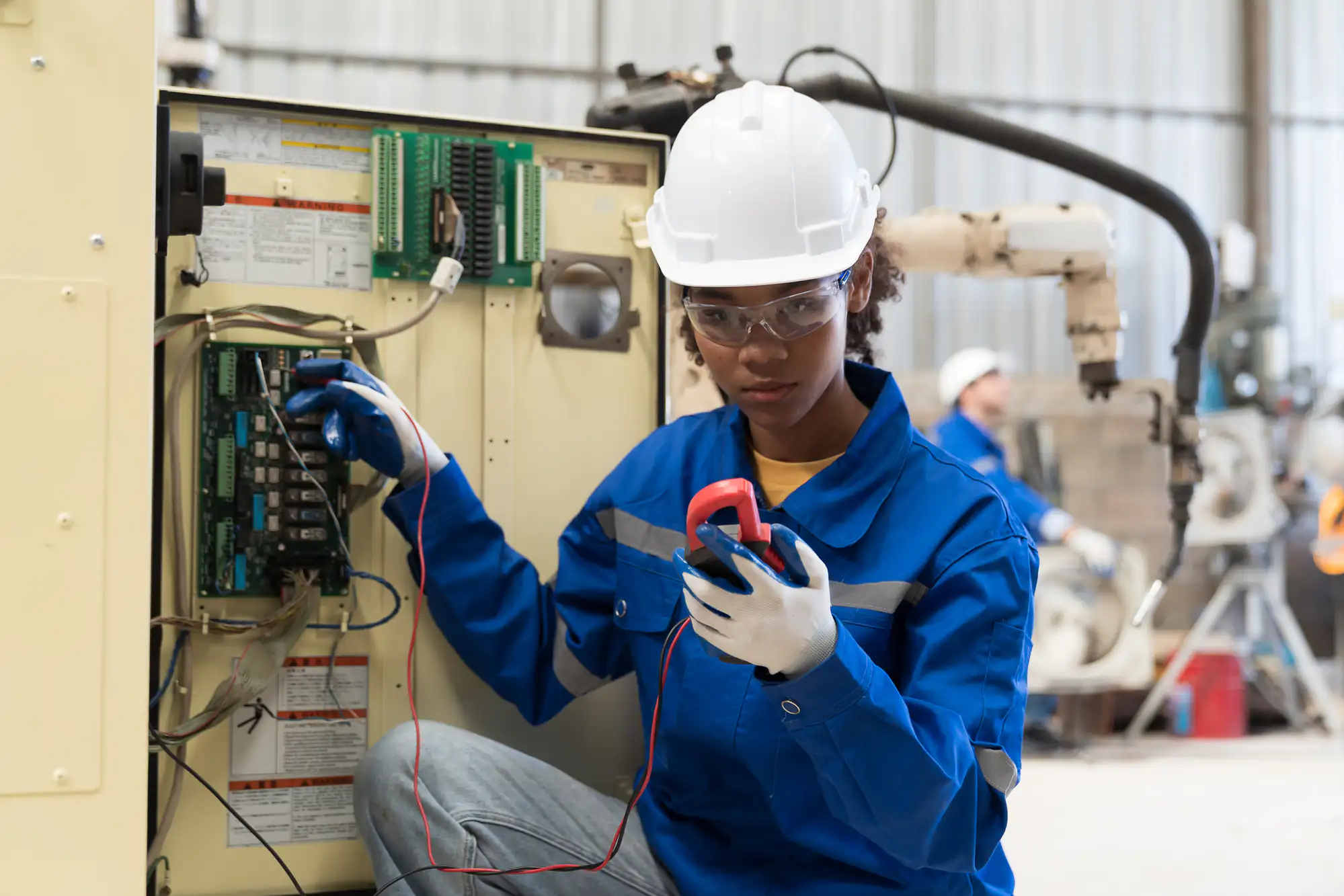 An electrician Brooklyn, NY, wearing safety gear, uses a multimeter to check electrical components inside an industrial control panel in a workshop or factory setting.