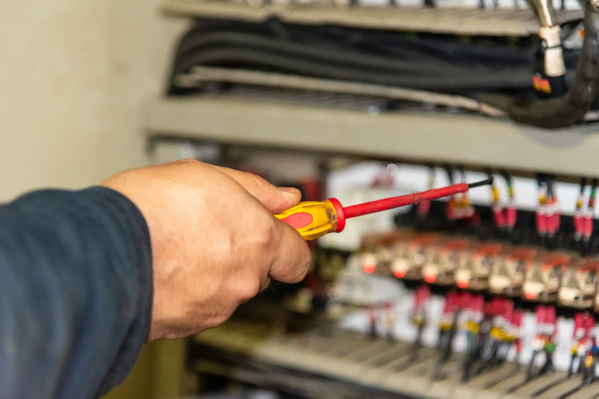A person&rsquo;s hand holds a red and yellow screwdriver, working on a panel filled with electrical wires and components&mdash;capturing the skilled touch of an electrician in Brooklyn, NY. The focus remains on the hand and tool, with the electrical panel slightly blurred behind.