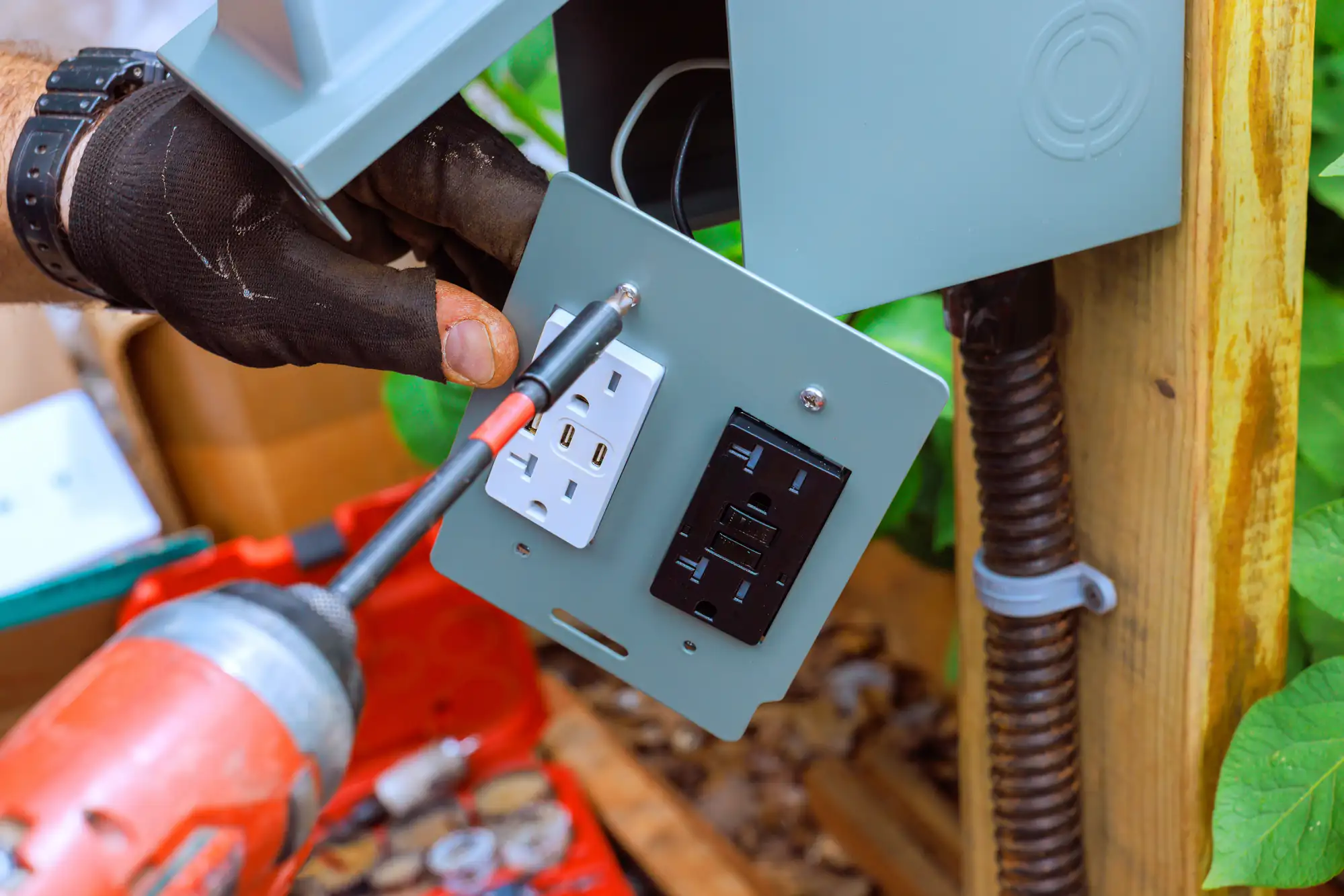 A gloved hand uses a red power drill to install an outdoor electrical outlet panel with standard and GFCI outlets on a wooden post, as an electrician Brooklyn expertly works in a leafy NY setting surrounded by tools.