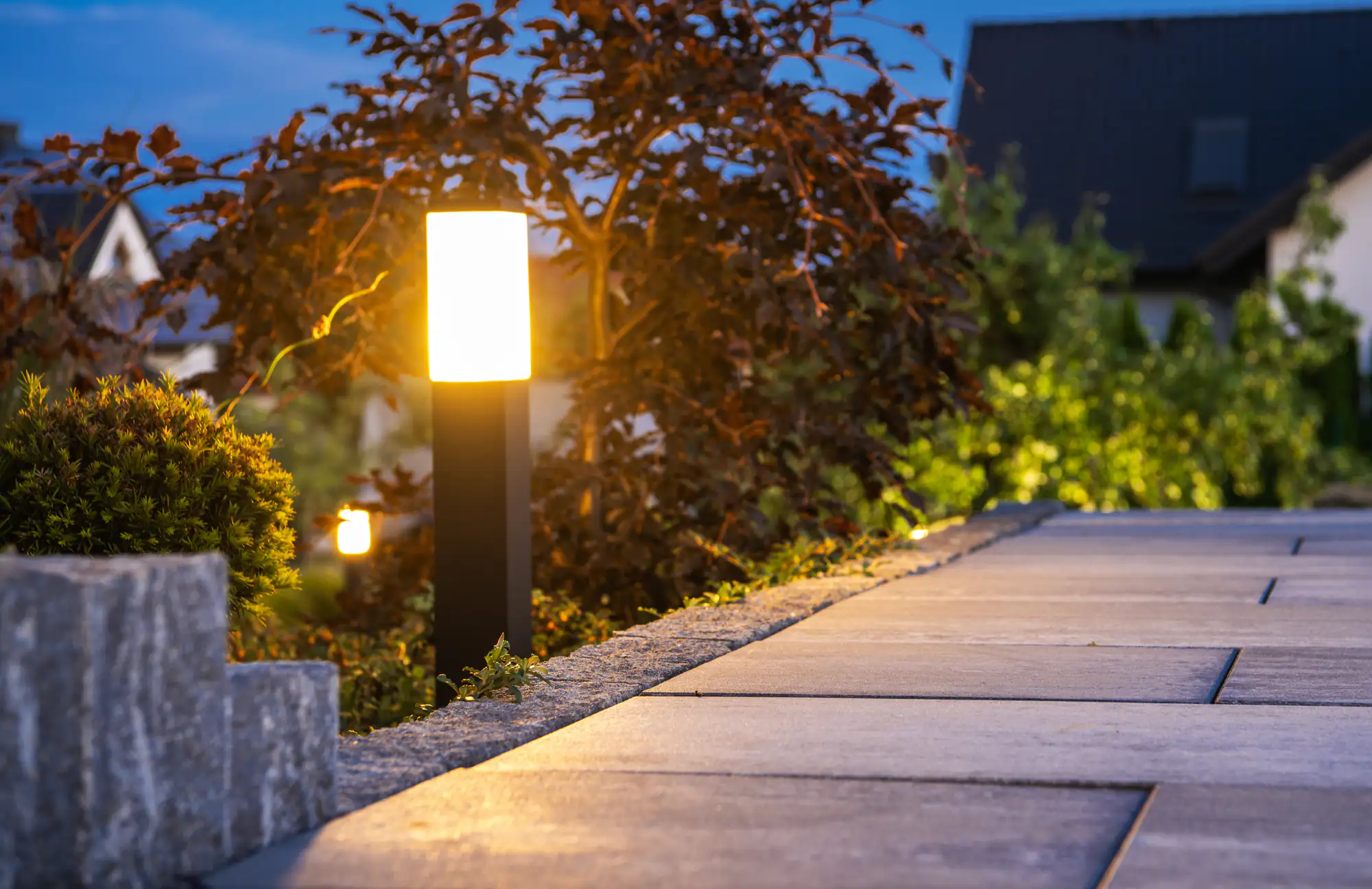 A glowing garden path light, expertly installed by an electrician in Brooklyn, NY, illuminates a stone walkway bordered by shrubs and trees in the evening, with houses softly blurred in the background.