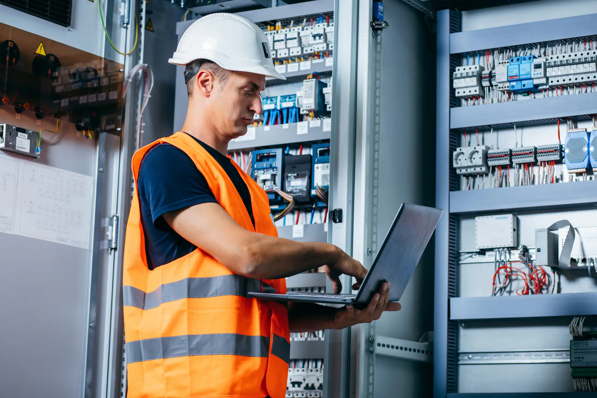 An electrician in Brooklyn, NY, wearing a white hard hat and orange safety vest, stands before open electrical panels, using a laptop to inspect or manage the equipment.