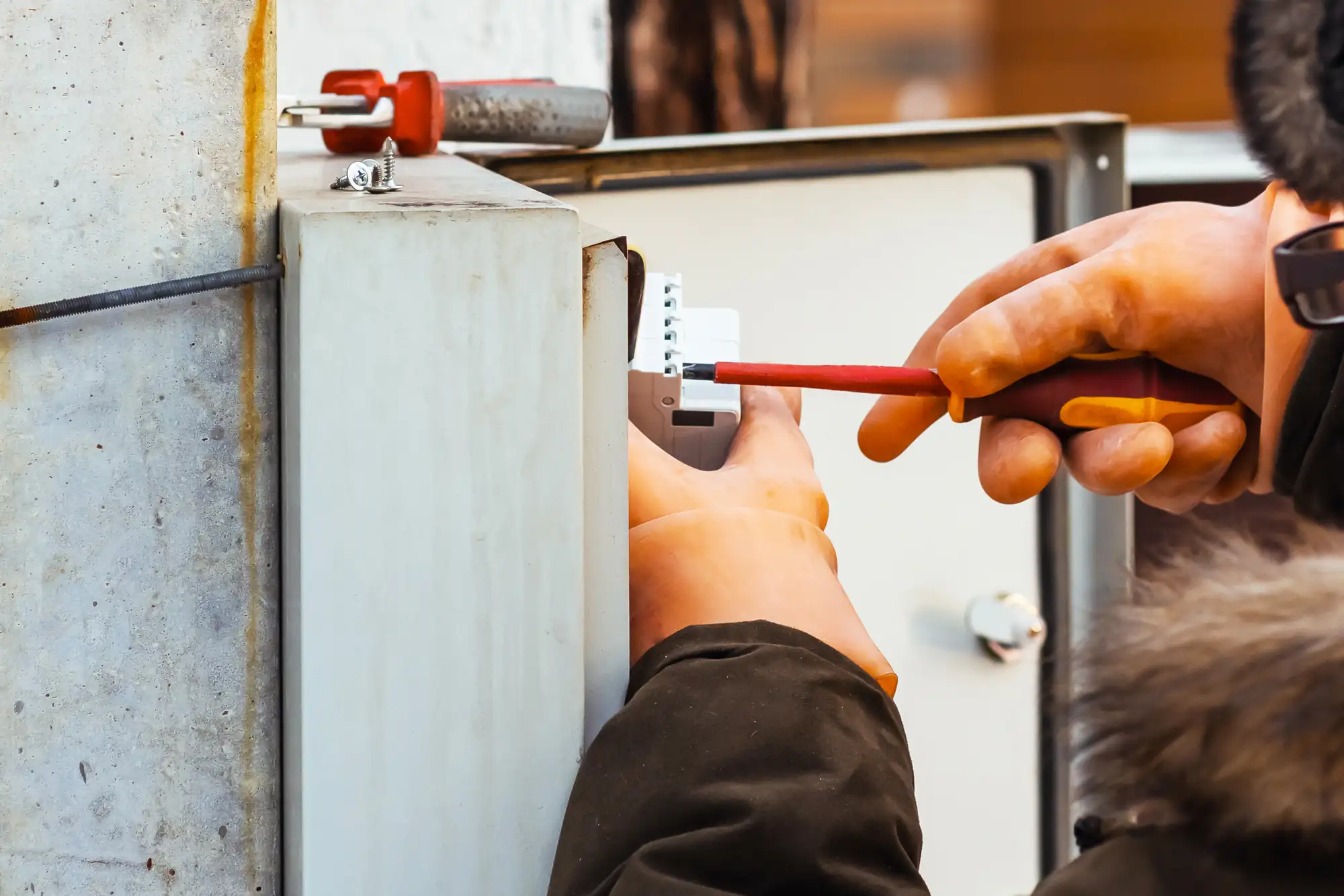 An electrician in Brooklyn, NY, wearing protective gloves, uses a screwdriver to connect or adjust a component inside an electrical panel mounted on the wall.