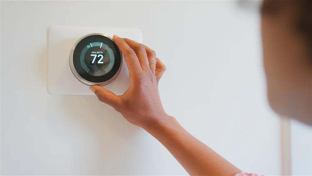 A person adjusts a smart thermostat on a wall, setting the temperature to 72 degrees Fahrenheit. The display reads "cool set to 72.