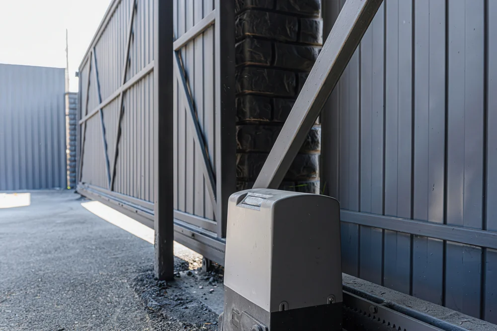 A close-up view of an automatic sliding metal gate with a motorized opener installed at the base, set on a paved driveway with metal fencing in the background.