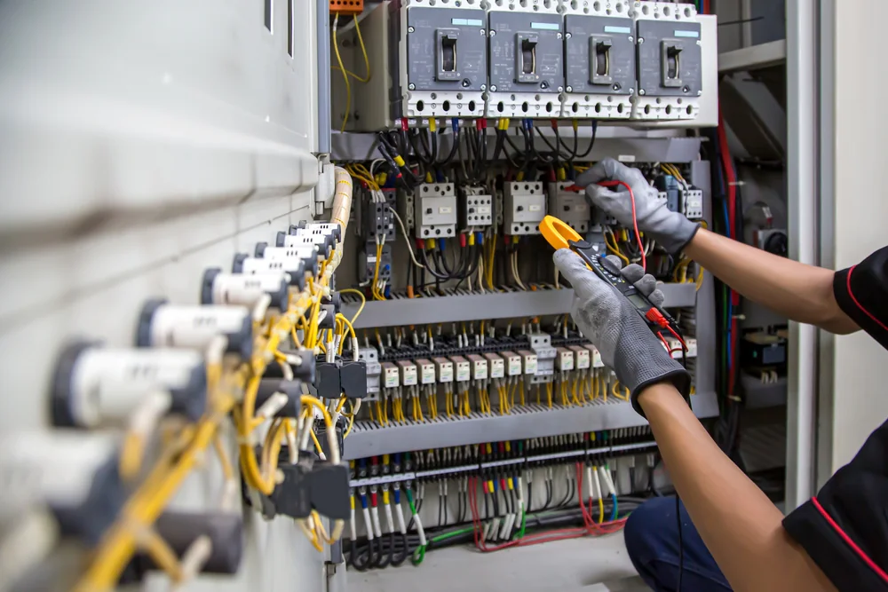 An electrician wearing gloves uses a clamp meter to test electrical connections inside an open control panel with numerous wires, switches, and circuit breakers.