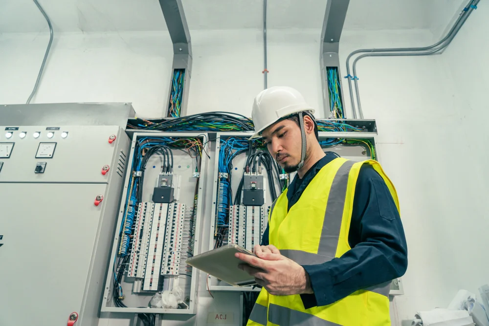 A worker in a safety vest and hard hat uses a tablet while standing in front of electrical control panels with exposed wiring inside an industrial facility.