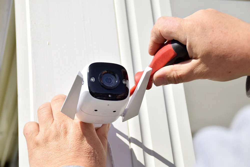 A person uses a screwdriver to install a security camera on a white wall or frame, securing it in place with a bracket.