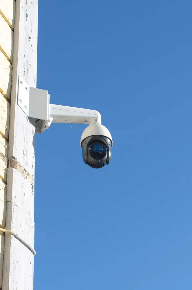 A white security camera mounted on a wooden wall faces outward against a clear blue sky.