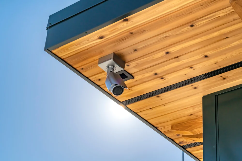 A security camera is mounted on a wooden ceiling beneath the edge of a modern building roof, with a clear blue sky in the background.