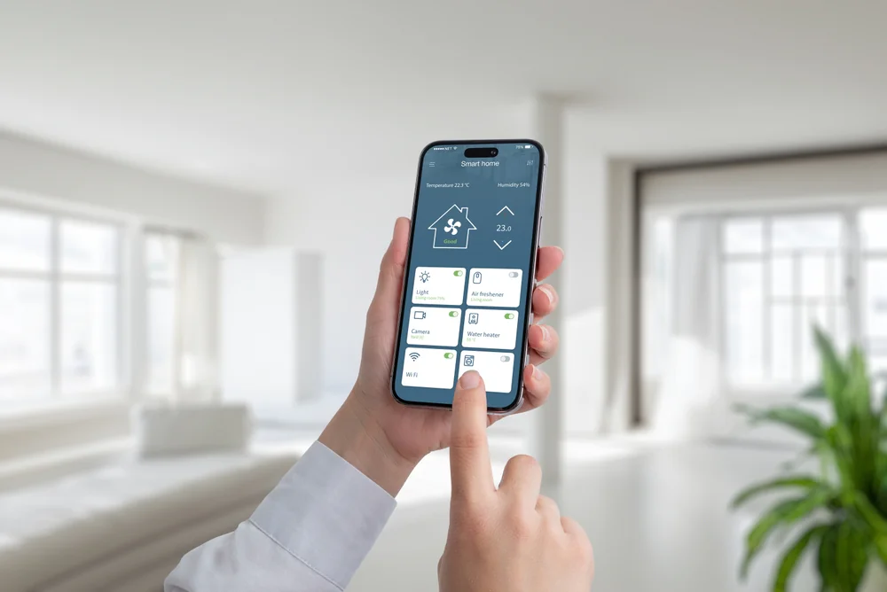 A person uses a smartphone to control smart home devices in a bright, modern living room. The phone screen displays options like lights, temperature, and security settings.
