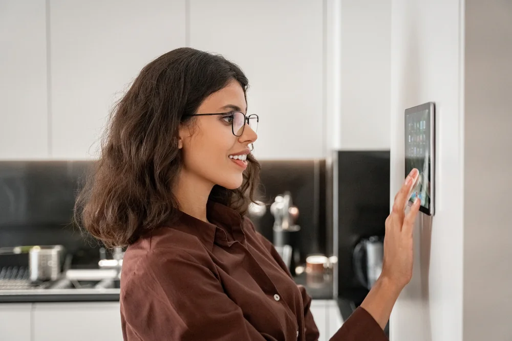 A woman with glasses and shoulder-length hair uses a smart home control panel mounted on a wall in a modern kitchen. She is smiling and wearing a brown shirt.