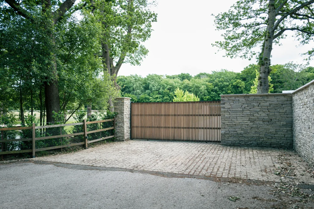 A closed wooden gate with vertical slats is set between two stone walls, surrounded by trees and greenery, with a wooden fence and paved driveway in the foreground.